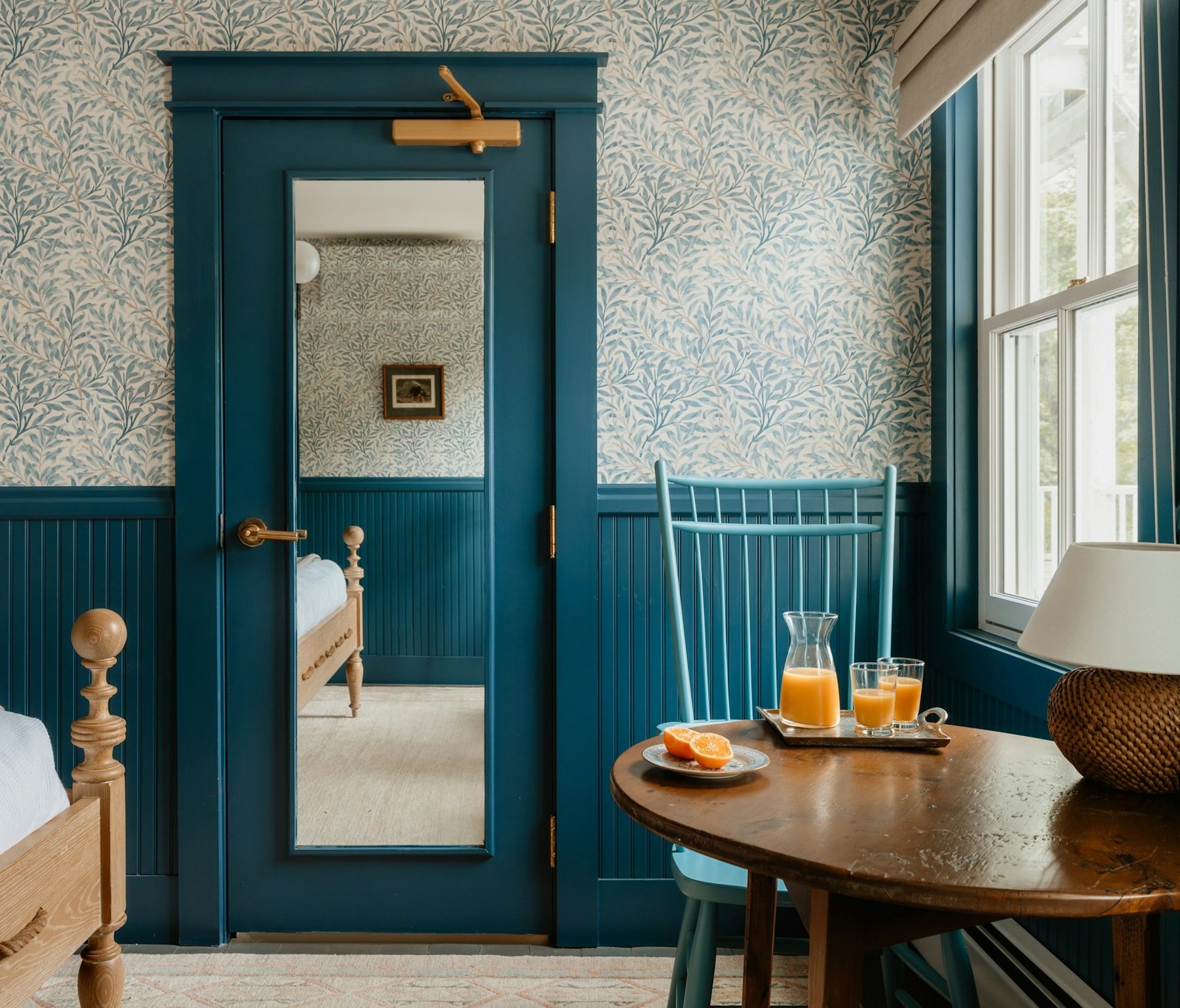 Bedroom papered in a blue-grey willow-leaf wallpaper above deep teal beadboard wainscoting, with a teal door, a window and a round wooden side table