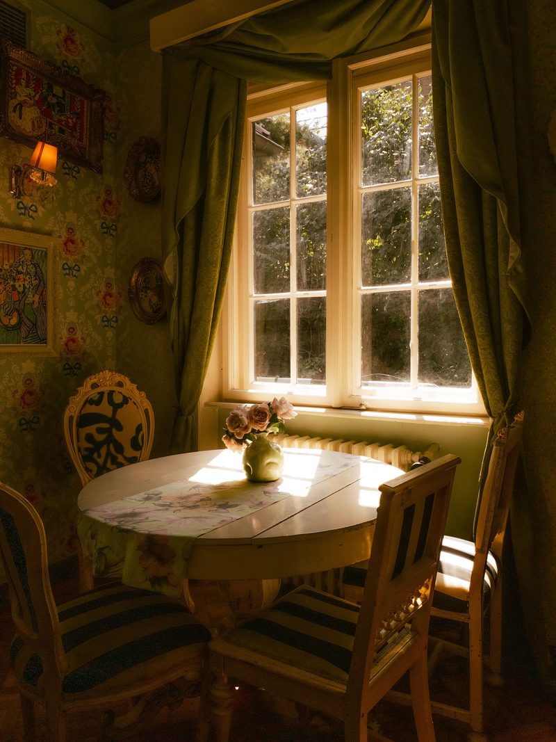 Sunlit breakfast nook with a traditional floral wallpaper, a round table and wooden chairs beside a window with long curtains
