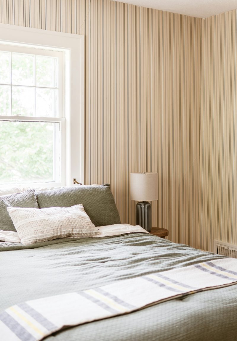 Quiet bedroom papered in a fine cream and taupe vertical stripe, with a double-hung window, sage bedding and a ribbed table lamp