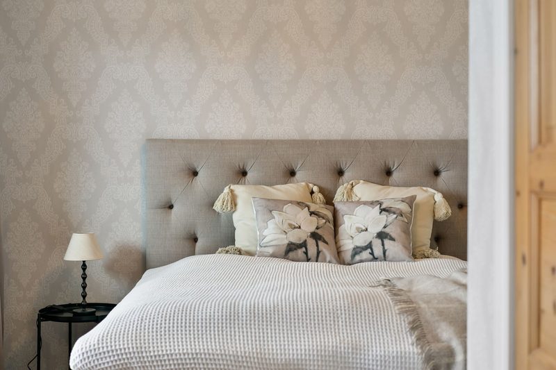 Bedroom with a soft neutral damask wallpaper behind a tufted linen headboard, white bedding and a small black side table